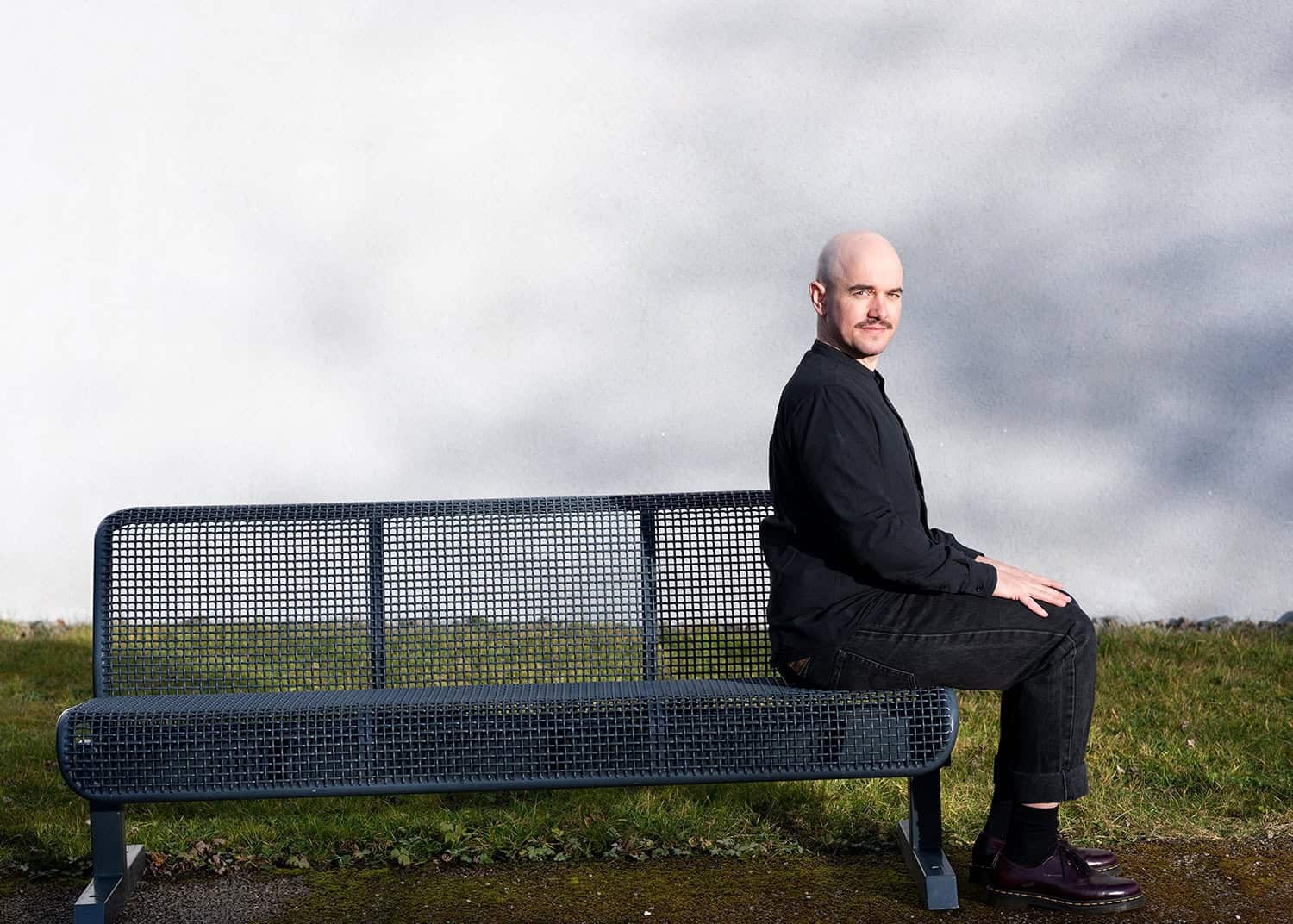 Renato Kaiser sitzt auf einer Bank bei Gleis21, Blick nach rechts, grauer Himmel.